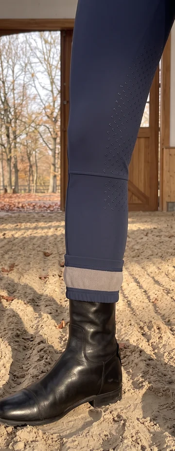 Umgeschlagener Saum einer Navy Winterreithose mit Fleecefutter über dem Reitstiefel, Herbstlaub auf dem Hallensand – KI-generierte Illustration